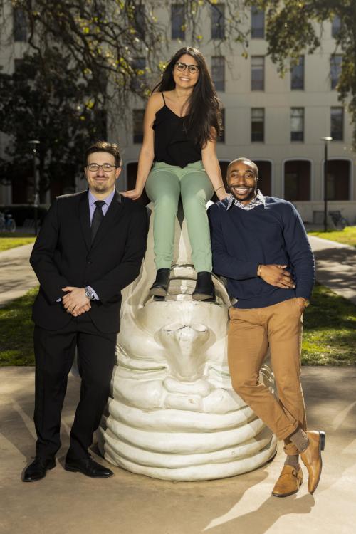 Two students stand on either side of an Egghead sculpture and one sits on top of the Egghead