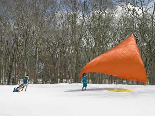 Image of two people standing in a snowy field with dark woods in background holding a large cone shaped orange fabric filled wiith air