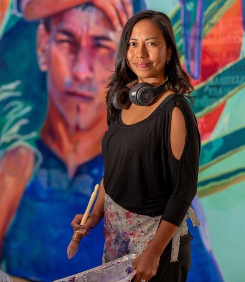 photo of women in black top holding paint brush in front of a large painting with a person's head and colorful imaage