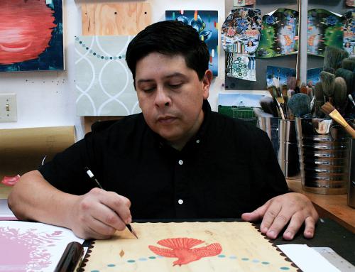 photo of a man in dark shirt, with dark hair drawing at a table. 