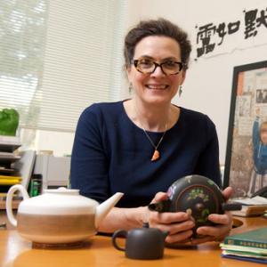 women with dark hair and glasses behind a desk holding tea pot 