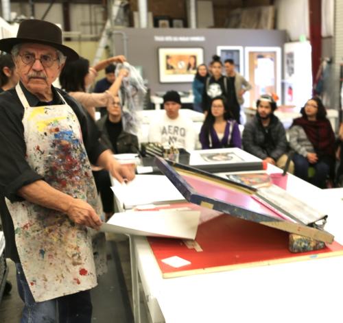 man with hat and ink coved apron demonstrating printmaking to students 