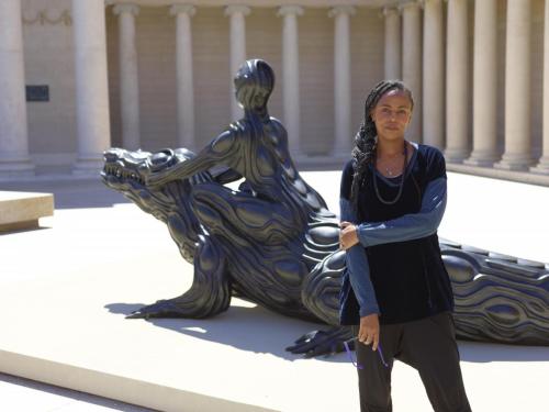 woman with dark skin standing in front of a large sculpture of woman and crocodil