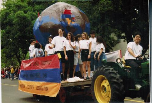 young man driving a green tractor pulling a wagon of float with nine young people wearing white shirts standing in front of a large globe. 