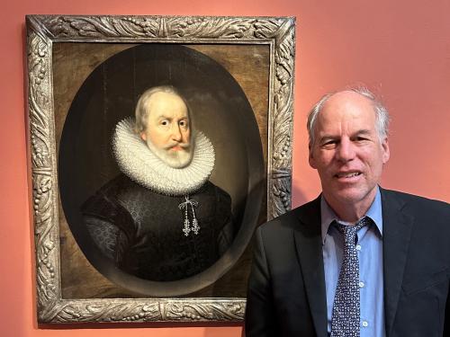 man in dark sport coat with blue shirt and dark blue tie standing next to a painting of a man's head and shoulder, with large white ruff and black jacket. 
