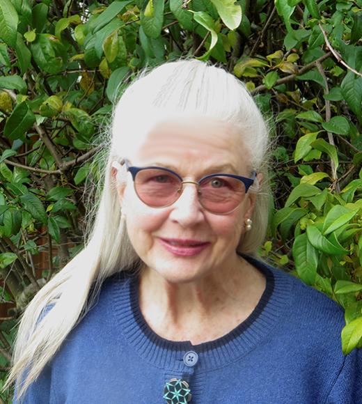 Smiling older woman with transition-lens glasses, long silver-gray hair pulled off her face, and blue top