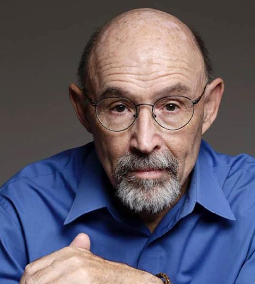 Headshot of older man with glasses, grey bead and bald head in blue collared shirt looking pensive