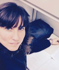 Young woman with dark hair and black jacket at her desk, looking up and over her right shoulder at camera