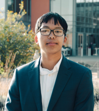 Asian American male student grinning, wearing eye glasses, white collared shirt and blue suit jacket, standing outside with campus building and greenery behind him