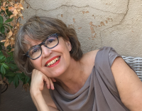 woman wearing gray loose blouse with chin length brown/gray hair , dark framed glasses, with head titled smiling