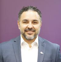 Man with dark hair with a little gray wearing light colored shirt and blue suit coat against a purple background 