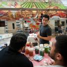 Photo of UC Davis muralist and two students, with mural in background and paints on table in foreground. 