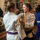 Annaliese Franz, smiling, holds back of fist near another woman's face while grabbing her lapel in a karate class.s
