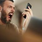 Man with beard looking outraged by what he sees on cell phone in his upraised hand.