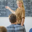 Blond female teacher, smiling at students in classroom, gestures at equations on blackboard