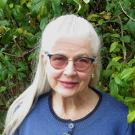Smiling older woman with transition-lens glasses, long silver-gray hair pulled off her face, and blue top