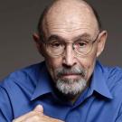 Headshot of older man with glasses, grey bead and bald head in blue collared shirt looking pensive