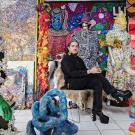 Photo of man with dark hair and black clothing sitting in a chair surrounded by art that is brightly colored 