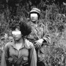 black and white photo of two women their eyes blanked out by white bars in military fatigues holding guns.