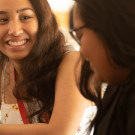 A young woman with brown hair and brown eyes looks left at a classmate and smiles.