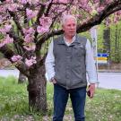 man wearing dark pants and gray vert standing outside in front of a tree with pink blossoms