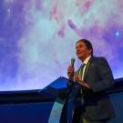 Manuel Calderón de la Barca Sánchez is holding a microphone and speaking in front of a large display of the universe behind him
