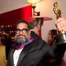 Bearded UC Davis alumnus dressed in tuxedo and a colorful scarf around his neck, holds up his Oscar backstage at the Academy Awards