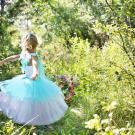 A little girl with a blond ponytail and a blue and white dress dances around a wooded area with flowers