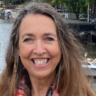 UC Davis psychology professor wearing bright floral scarf and smiling. Behind her is an Amsterdam canal, boats, a dock, bridges and a nearby tree-lined street
