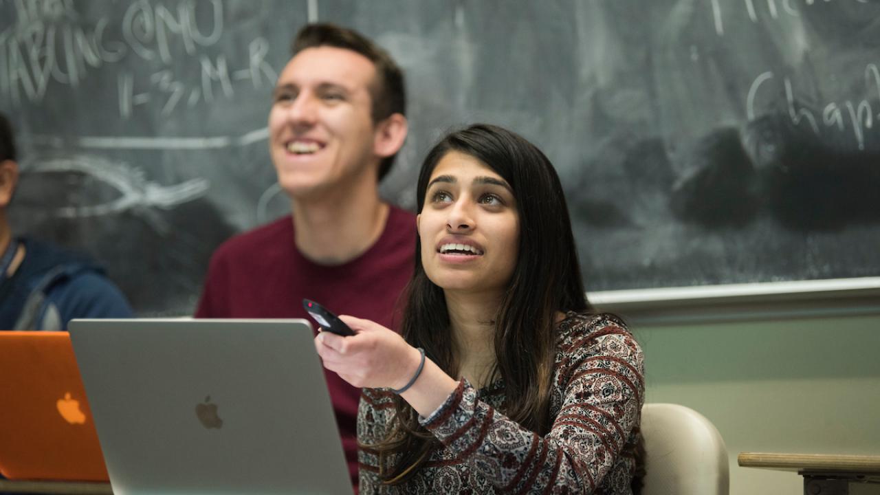 Female student speaking at laptop, male smiling behind, chalkboard with equations