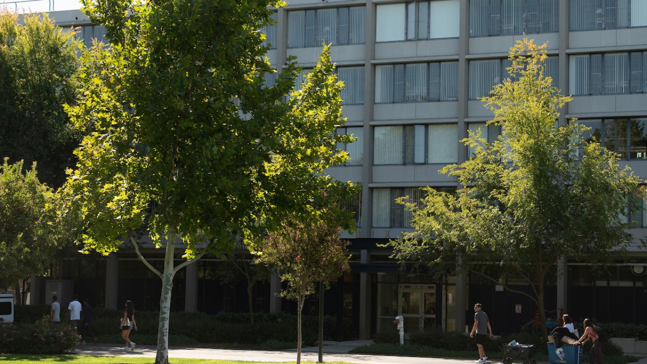Campus courtyard photo with green lawn, trees, and people sitting near a modern building