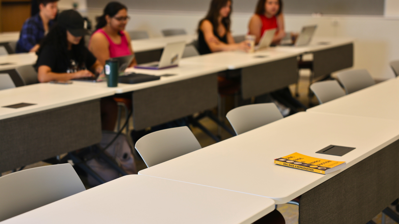 A photo focuses on a yellow book laying on the top of a table in a classroom at Aggie Square. Students in the Medical Humanities minor sit in the background, preparing for class to start.