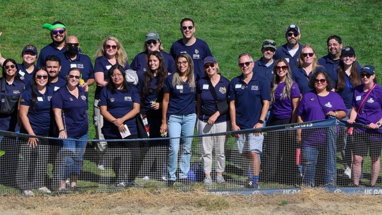 Group in navy and purple shirts standing behind chain-link fence on a grassy field, smiling