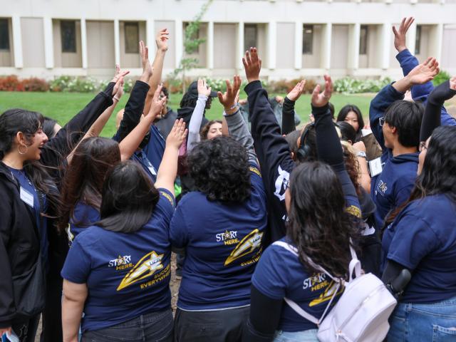 Group of students in navy shirts raising hands together in a campus courtyard