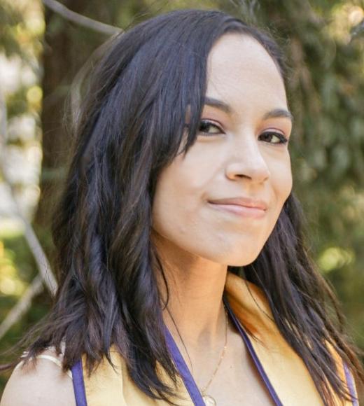 Headshot of young person with straight black hair just past shoulders, grinning and wearing gold graduation stole, green trees in sun in background