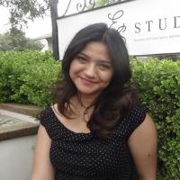 Smiling woman with long dark hair in a black polka-dot dress seated outdoors by greenery and sign