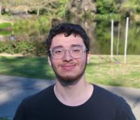 Smiling young man with glasses standing outdoors near a pond and greenery.