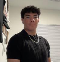 Young man with dark curly hair in black shirt wearing AirPods, standing in a bedroom