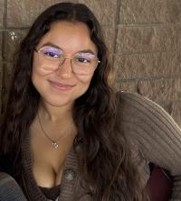 Smiling woman with long dark hair and glasses in brown cardigan, seated against textured wall