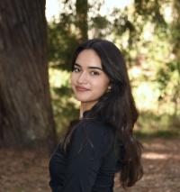 Young woman with long dark hair in black top, smiling, turned toward camera in sunlit forest.