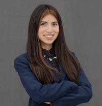Smiling young woman with long brown hair in navy blazer, arms crossed against gray background