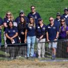 Group in navy and purple shirts standing behind chain-link fence on a grassy field, smiling