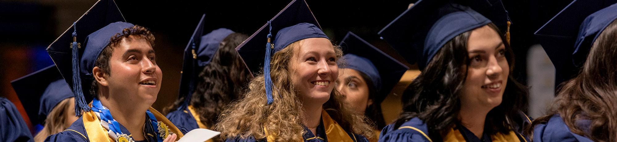 A group of 2021 UC Davis graduates wearing masks and caps, facing away from the camera, against a background of green leaves